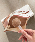 Hand holding a package of Juice Plus+ Chocolate powder with a spoonful of powder visible.