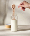 Person pouring a scoop of Juice Plus+ Complete powder into a glass of milk on a neutral background