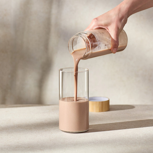 Person pouring a creamy liquid from a glass jar into another glass on a neutral background