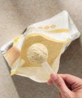 Hand holding a spoon with Juice Plus+  Complete powder over a package of the same powder on a neutral background