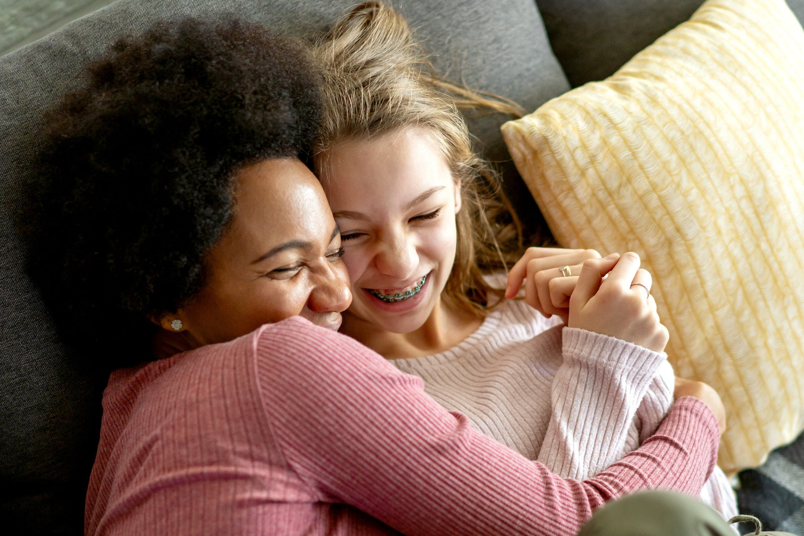 A close-up lifestyle shot of a woman and a smiling young girl hugging on a gray couch, with a yellow striped pillow in the background.