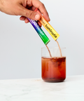 Hand pouring a Juice Plus+ Luminate and Superfood Powder into a glass of iced coffee on a white background