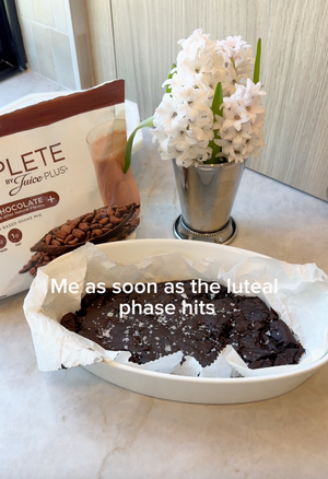 Brownie in a dish with a package of Complete by Juice Plus+ and a vase of white flowers on a marble surface.