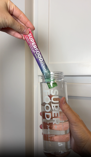 Hand holding a Superfood Paradise powder stick and a clear bottle labeled 'Superfood' against a white background.