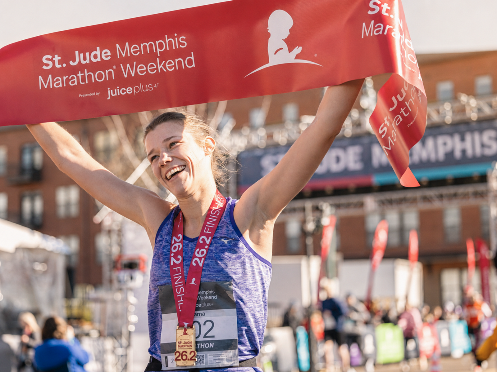 A smiling marathon runner holds up a red St. Jude Memphis Marathon Weekend banner with both arms raised, wearing a blue athletic tank top and a red race medal with a bronze finish.